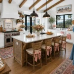 Bright kitchen with a rustic wood island, white cabinetry, woven bar stools, blush floral arrangements, coastal shell decor, black pendant lights, and ocean-view windows.